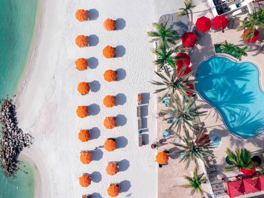 Vista aérea de una playa con filas de tumbonas y sombrillas, una piscina y palmeras.