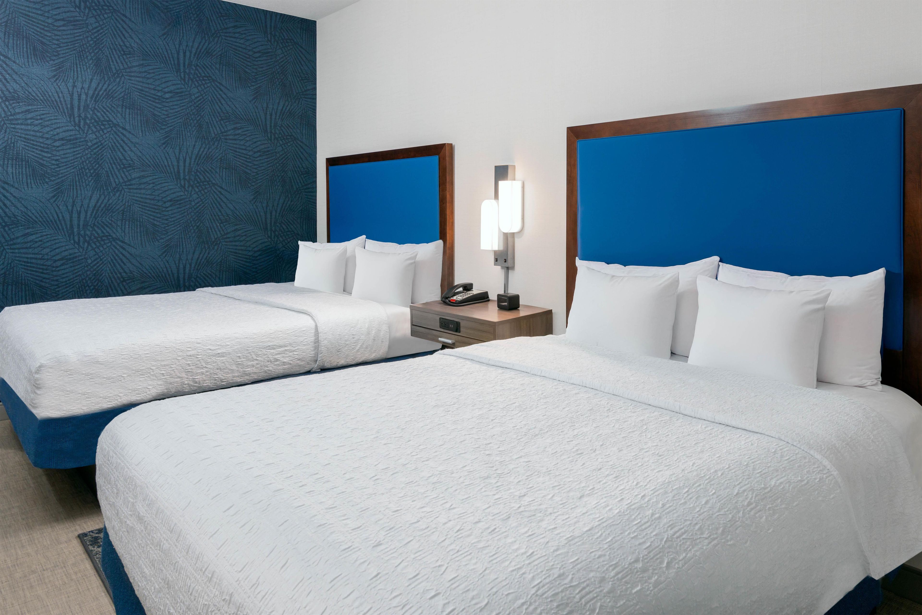A hotel room with two beds, a nightstand with a phone, and a wall-mounted light.