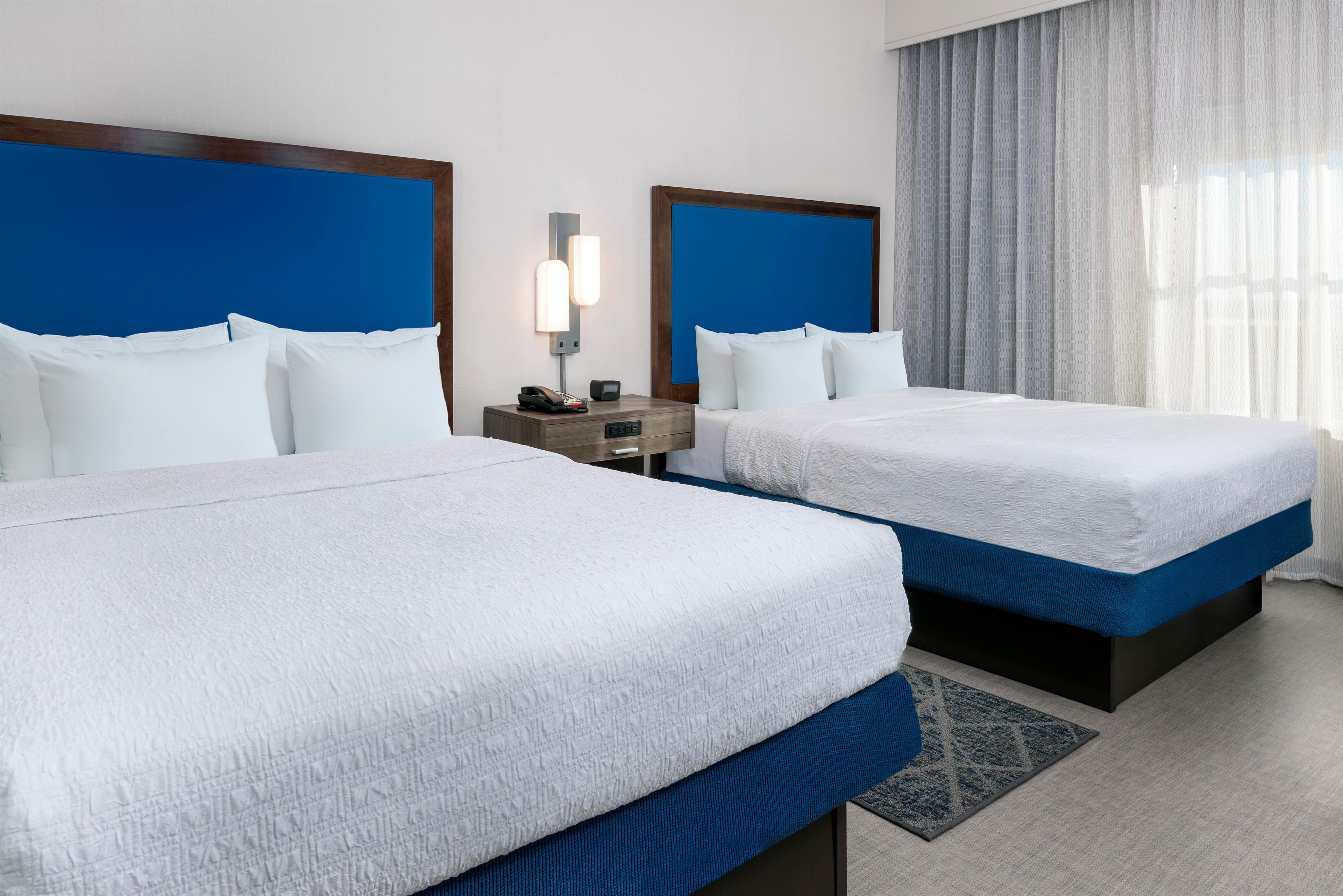 A hotel room with two beds, a nightstand with a phone and a small table, and a window with curtains.