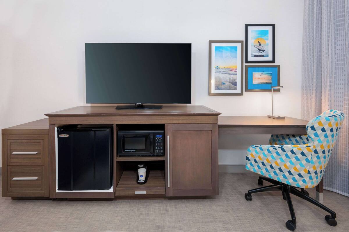 A compact hotel room with a built-in desk, a television, a microwave, and a small refrigerator. There is a chair with a geometric pattern and a framed picture on the wall.