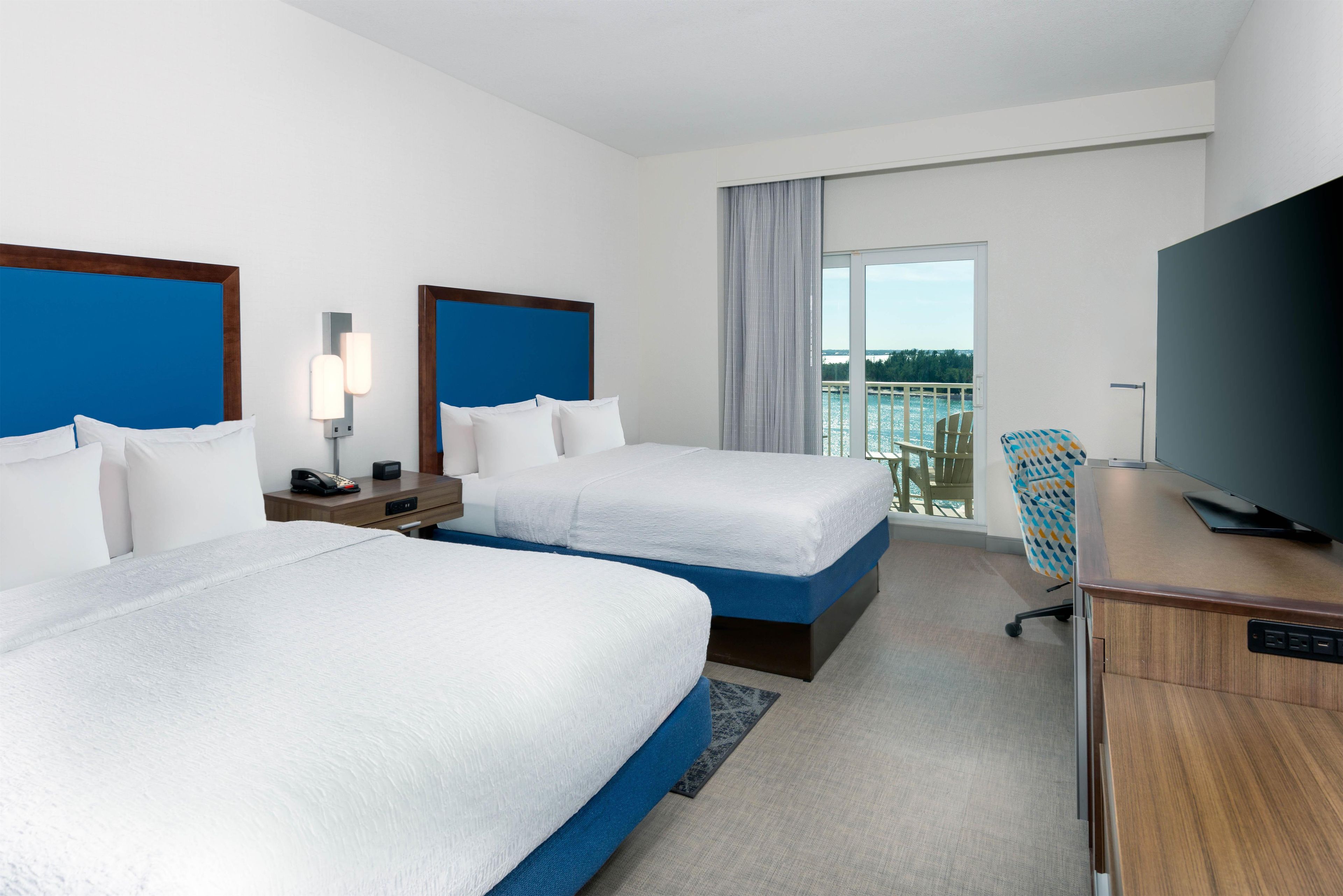 A hotel room with two beds, a desk, a television, and a balcony with a view.