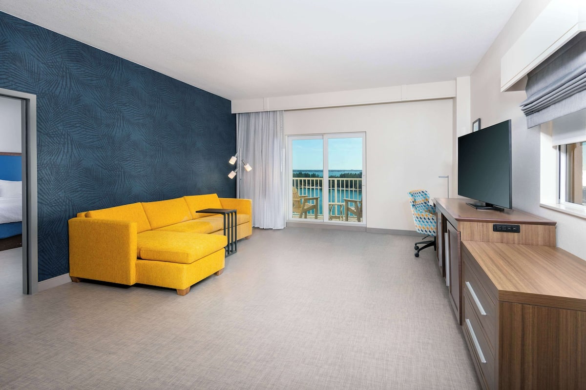 A modern hotel room with a yellow sofa, a TV on a wooden cabinet, and a balcony with a view.