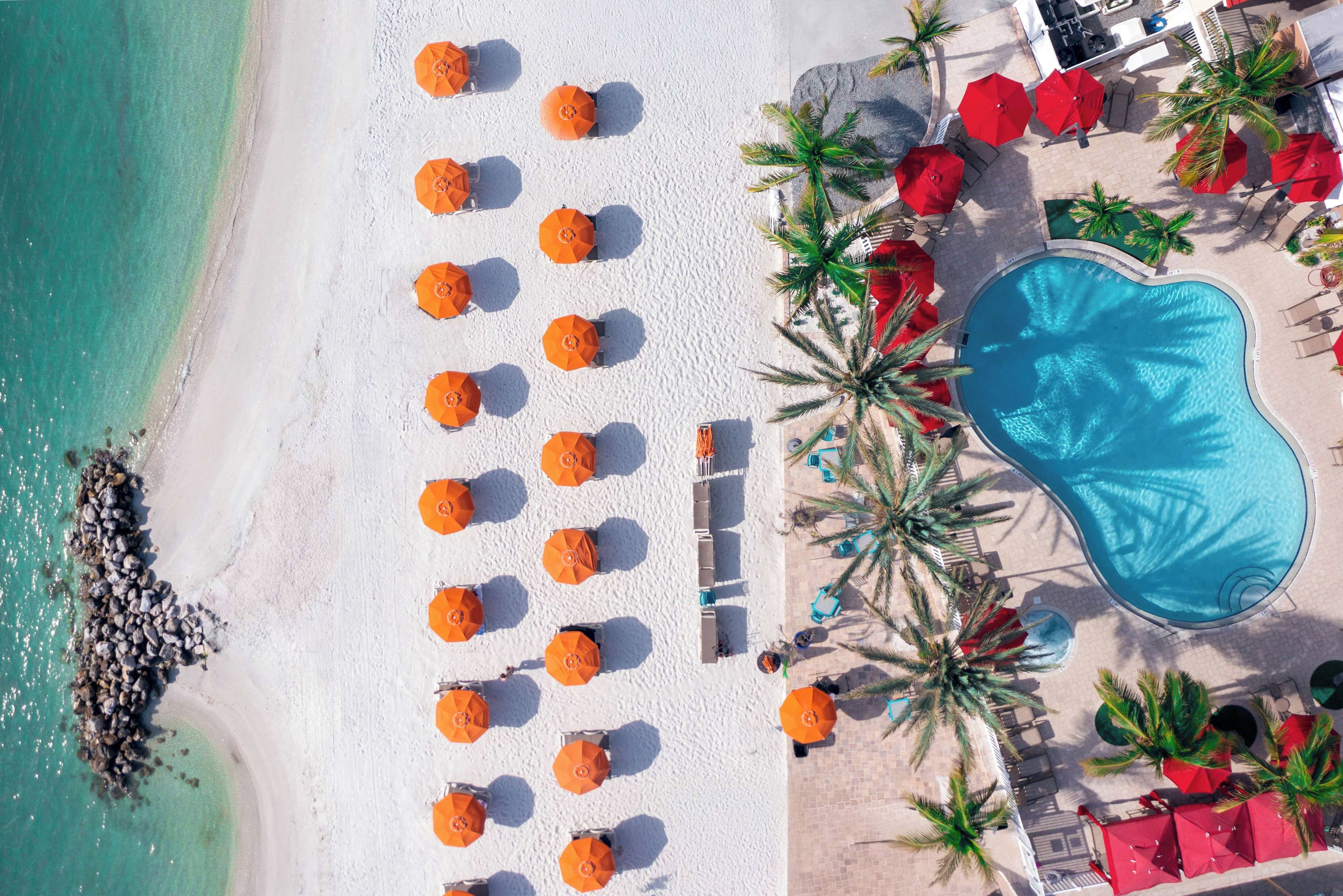 Vue aérienne d’une plage de sable blanc, avec une piscine et de nombreux parasols orange.