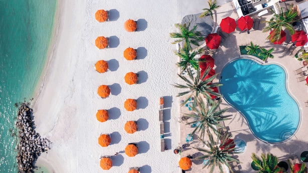 An aerial view of a beach with white sand, a swimming pool, and numerous orange umbrellas.