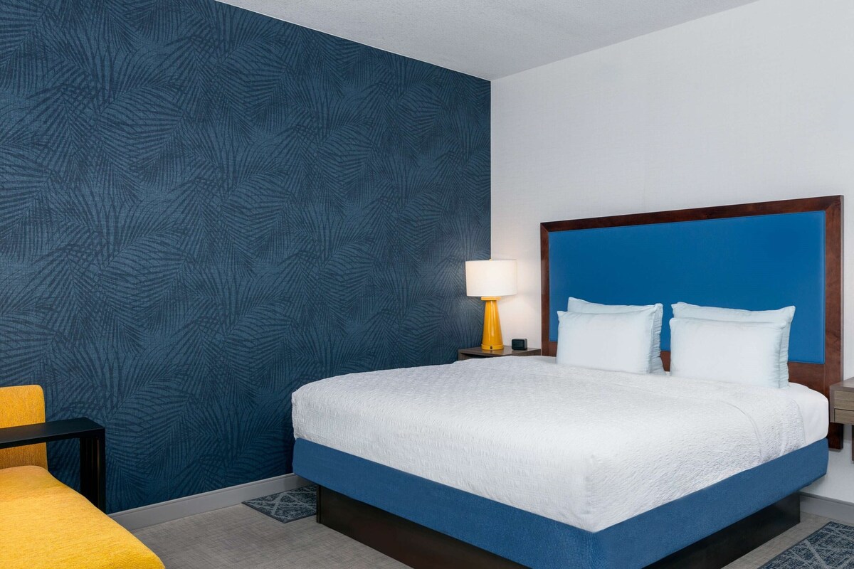 Ein Hotelzimmer mit einem großen Bett, einem Nachttisch mit Lampe, einem Sessel und einer Wand mit Muster.