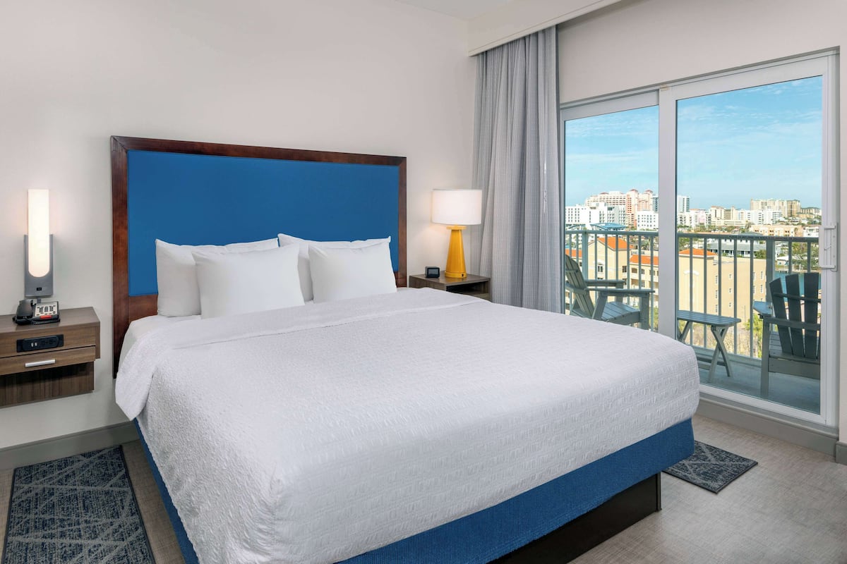 A hotel room with a large bed, a nightstand with a lamp, a view of the city through large windows, and a balcony with chairs.