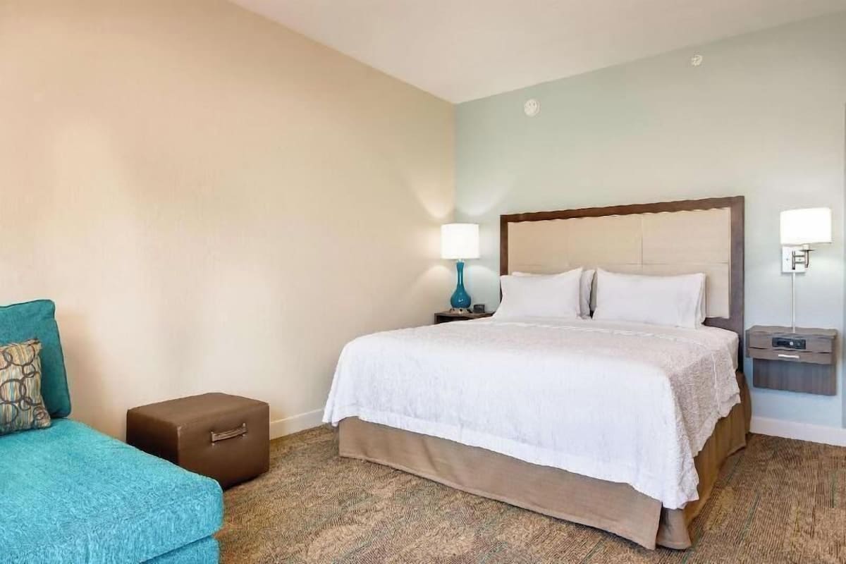 A hotel room with a bed, a nightstand, a lamp, a small table, and a suitcase.