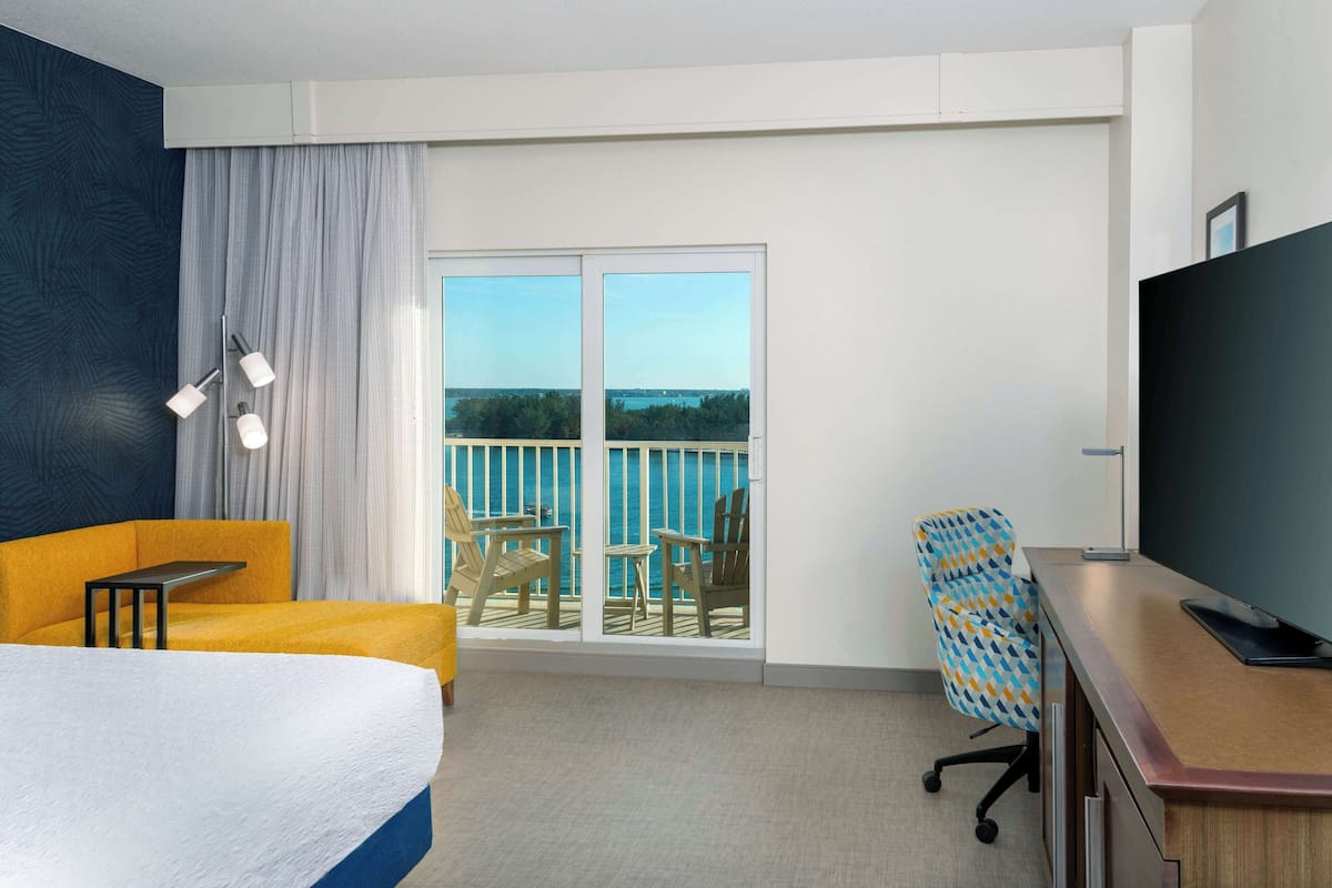A hotel room with a bed, a desk, a television, and a balcony with chairs and a view of the sea.