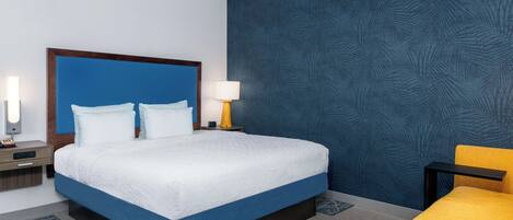 A hotel room with a large bed, a nightstand with a lamp, a small table, and a yellow chair.