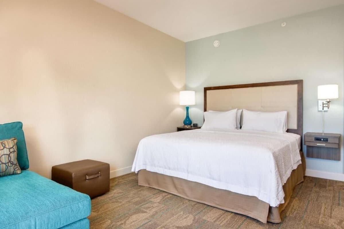 A hotel room with a bed, a small table, a nightstand, a lamp, a sofa, and a suitcase.