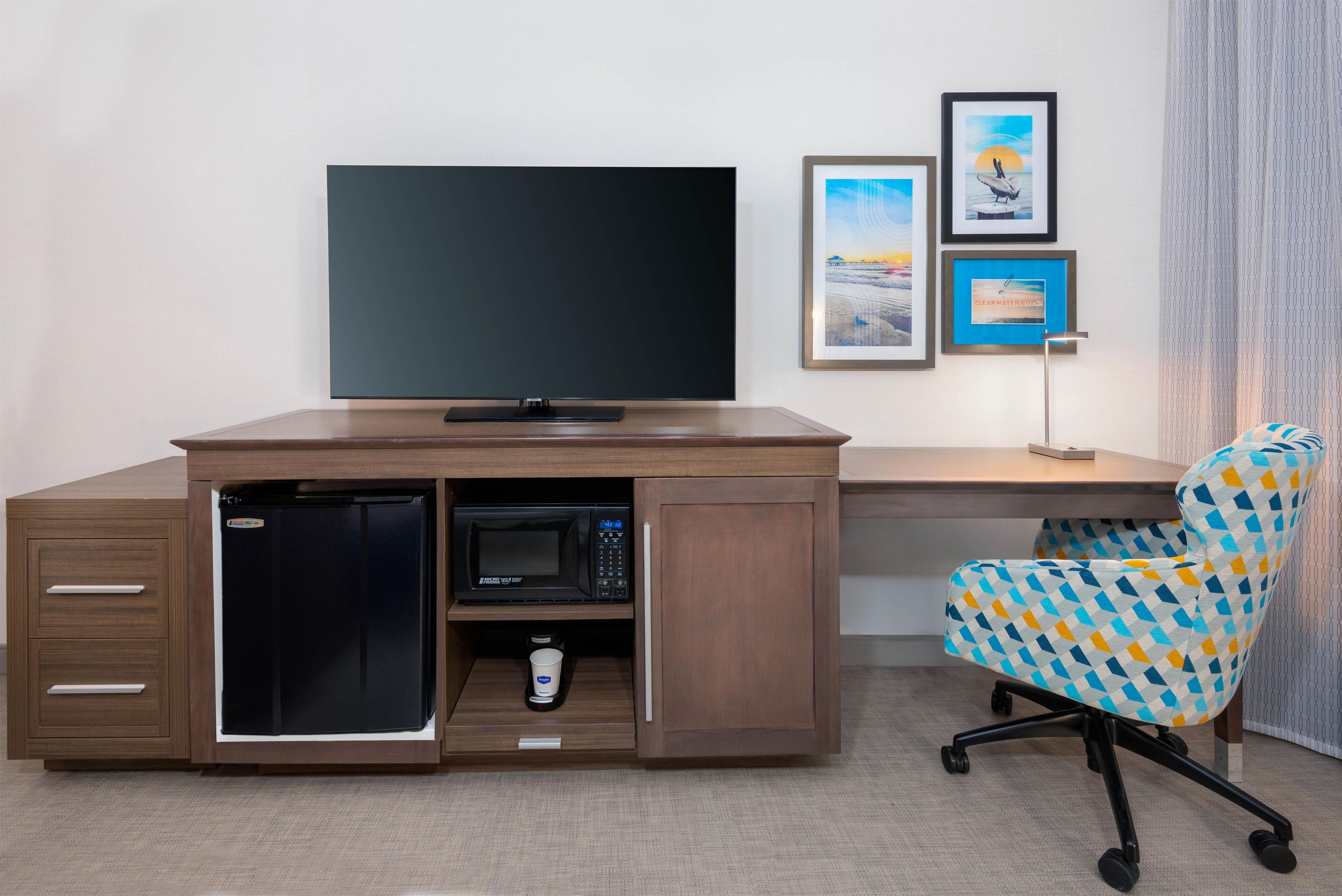 Une chambre d’hôtel compacte comprenant un bureau en bois, un four à micro-ondes encastré, un téléviseur à écran plat, un petit réfrigérateur, une chaise de bureau à motifs et des tableaux encadrés accrochés au mur.