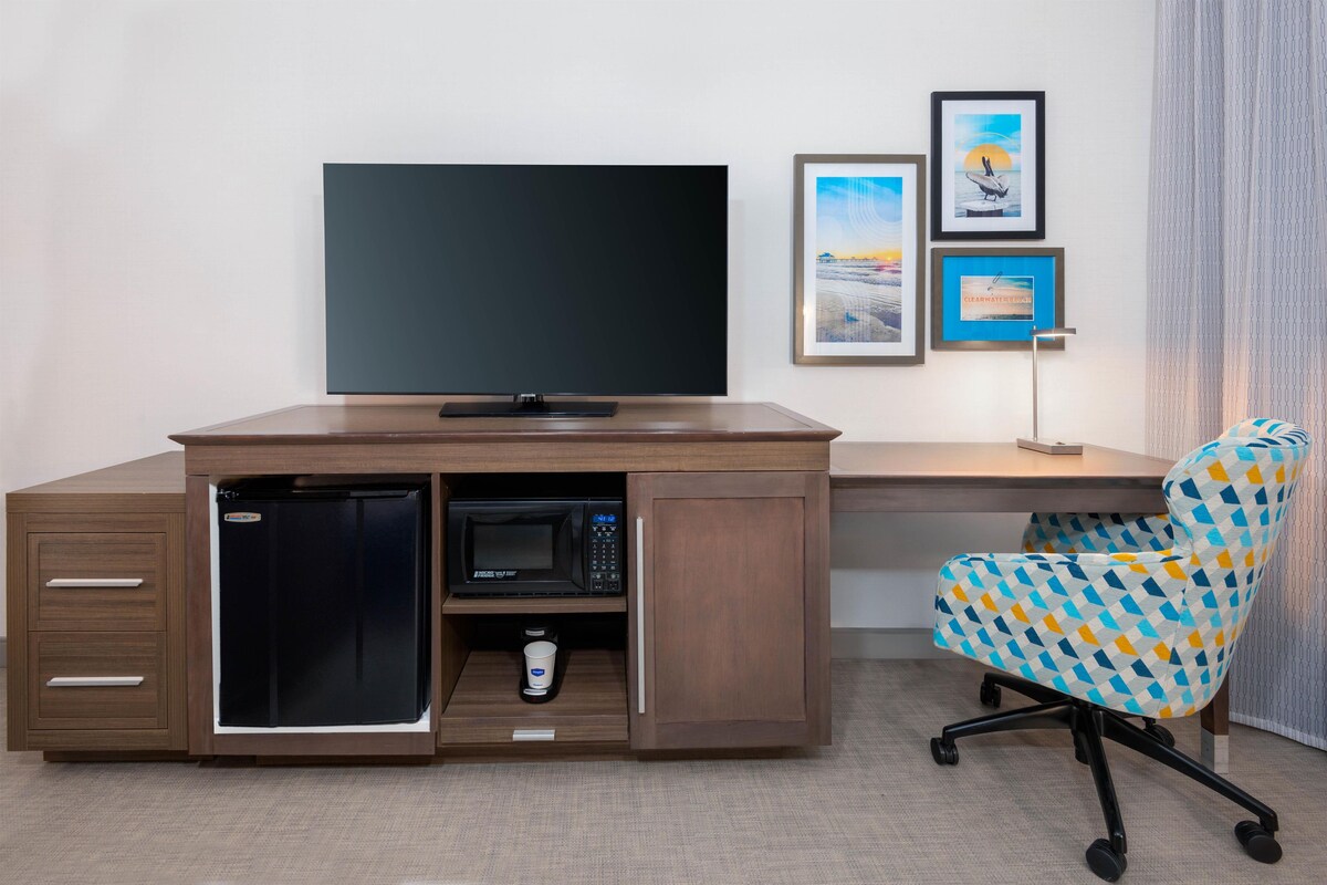 A compact hotel room with a wooden desk, a built-in microwave, a flat-screen TV, a small refrigerator, a patterned office chair, and framed pictures on the wall.