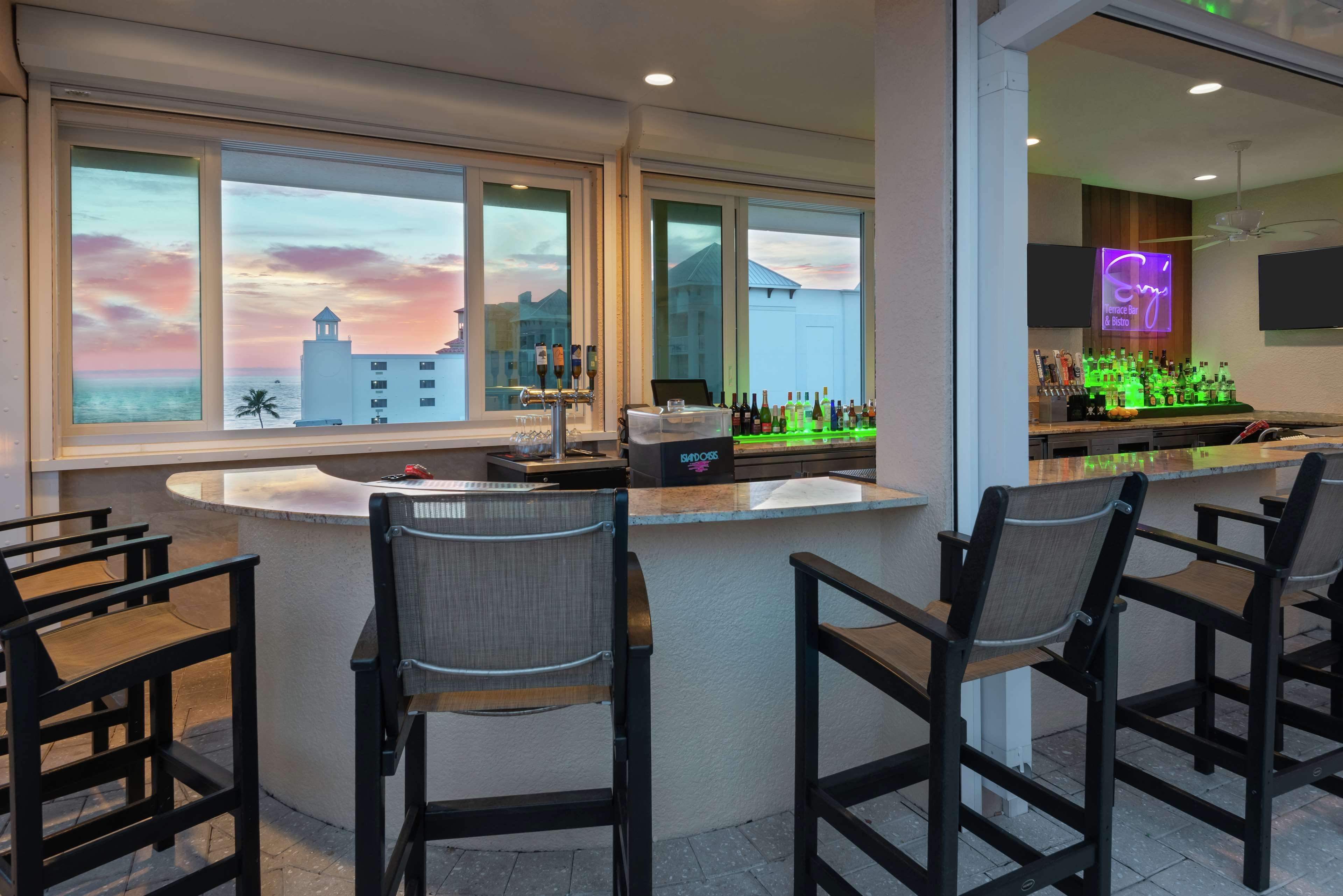 Eine Bar mit Blick auf einen Sonnenuntergang über dem Meer, Barhocker und einer Theke mit Flaschen.