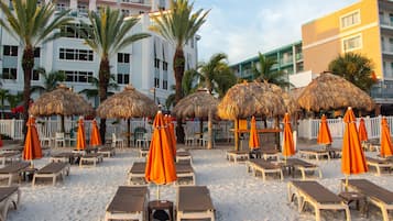 Une zone aménagée au bord de la plage, avec des chaises longues et des parasols en chaume, des palmiers et une clôture blanche.