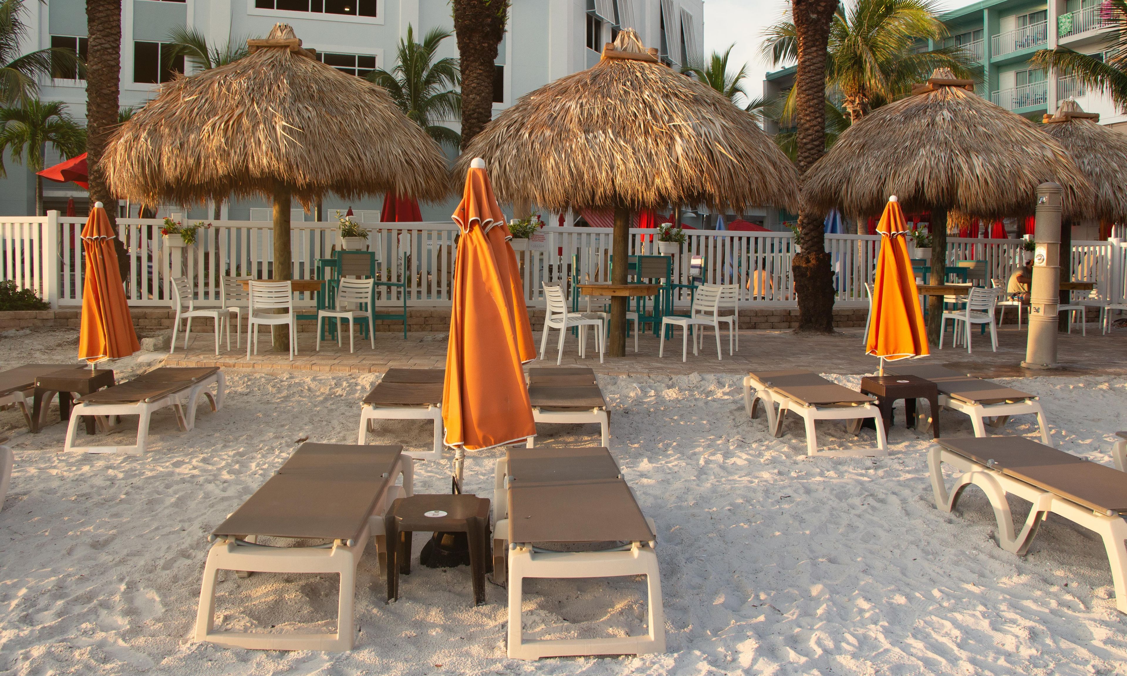 Een terras aan het strand met ligbedden en rieten parasols, omgeven door een witte hek en palmbomen.