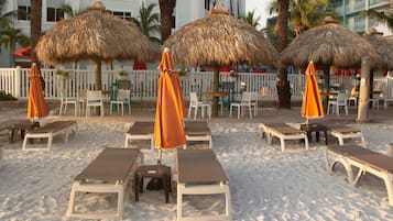 A beachside lounge area with sun loungers and thatched umbrellas, surrounded by a white fence and palm trees.