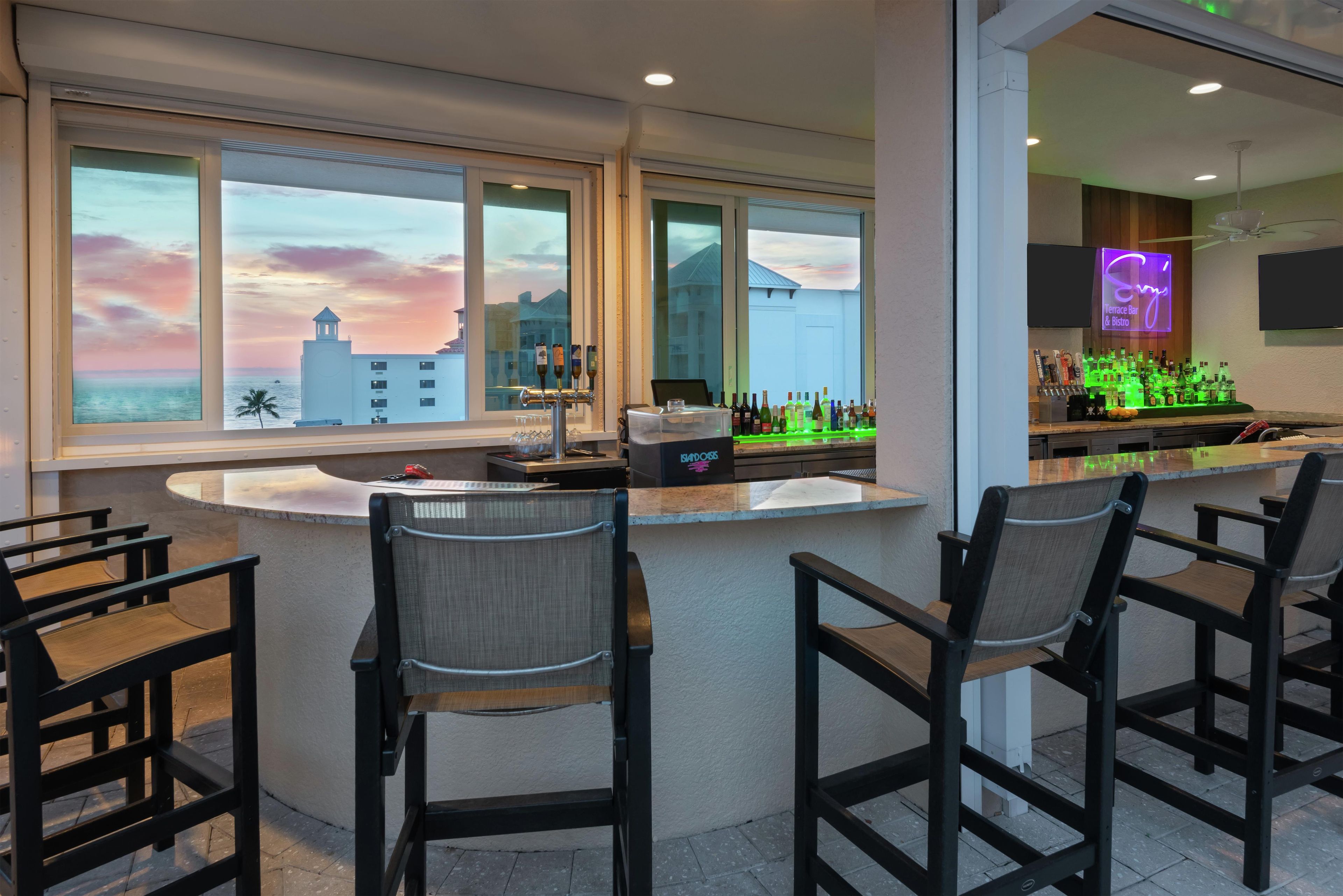 A bar area with a view of a sunset over a cityscape, bar stools, and a counter with bottles.