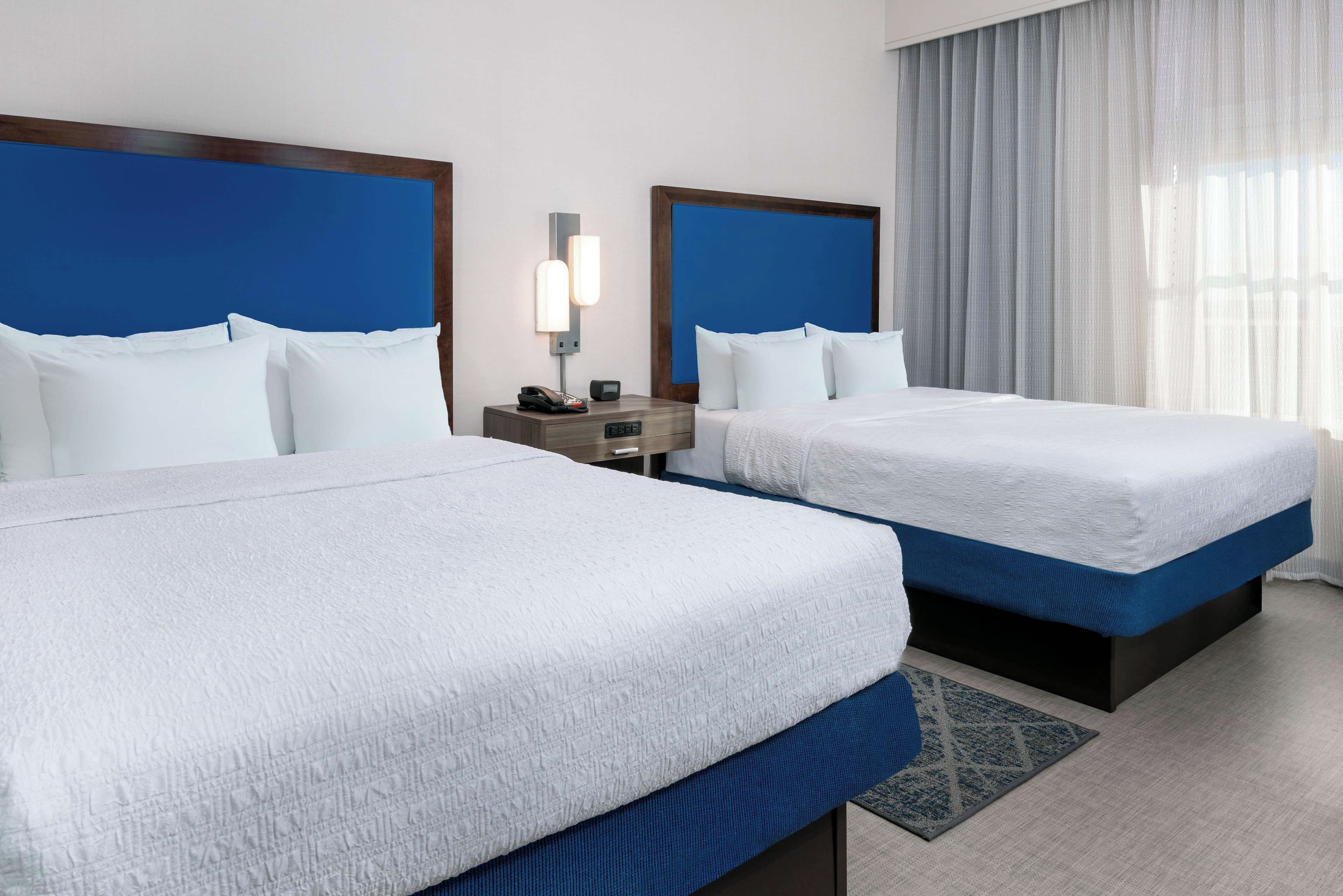 Une chambre d’hôtel avec deux lits, chacun doté de draps blancs et d’une tête de lit bleue. Une table de chevet avec une lampe et un téléphone se trouve entre les deux lits.