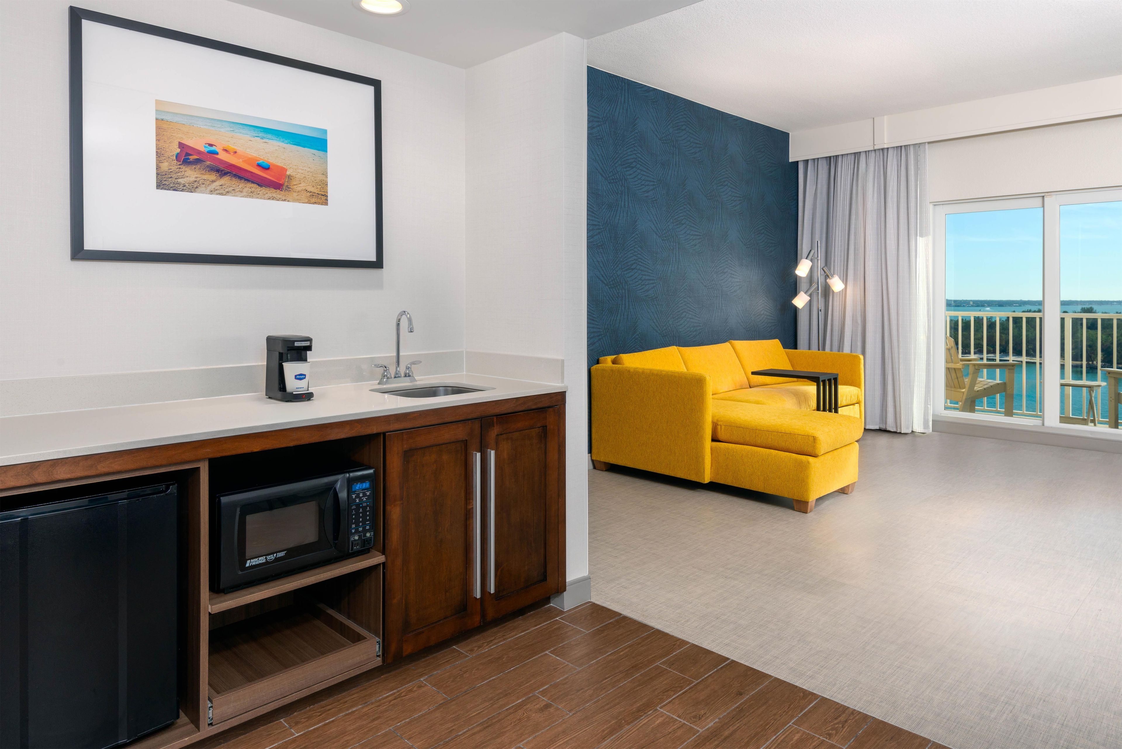 A modern hotel room with a kitchenette, a yellow sofa, a large window with a view, and a framed picture on the wall.