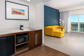 Habitación de hotel moderna con kitchenette, sofá amarillo, ventana grande con vistas y un cuadro enmarcado en la pared.