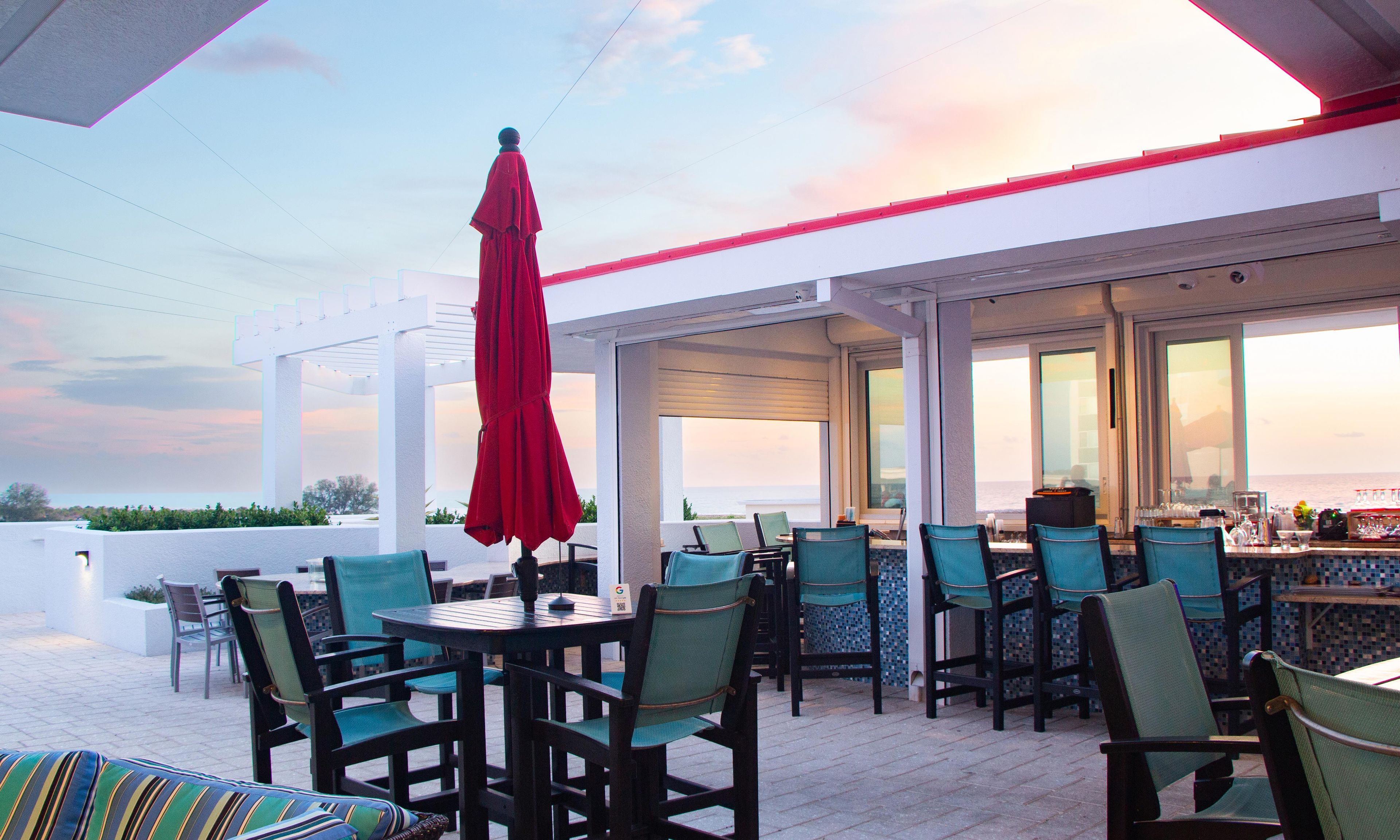 Un espace extérieur aménagé avec des tables et des chaises, un parasol rouge et une vue sur l’océan.