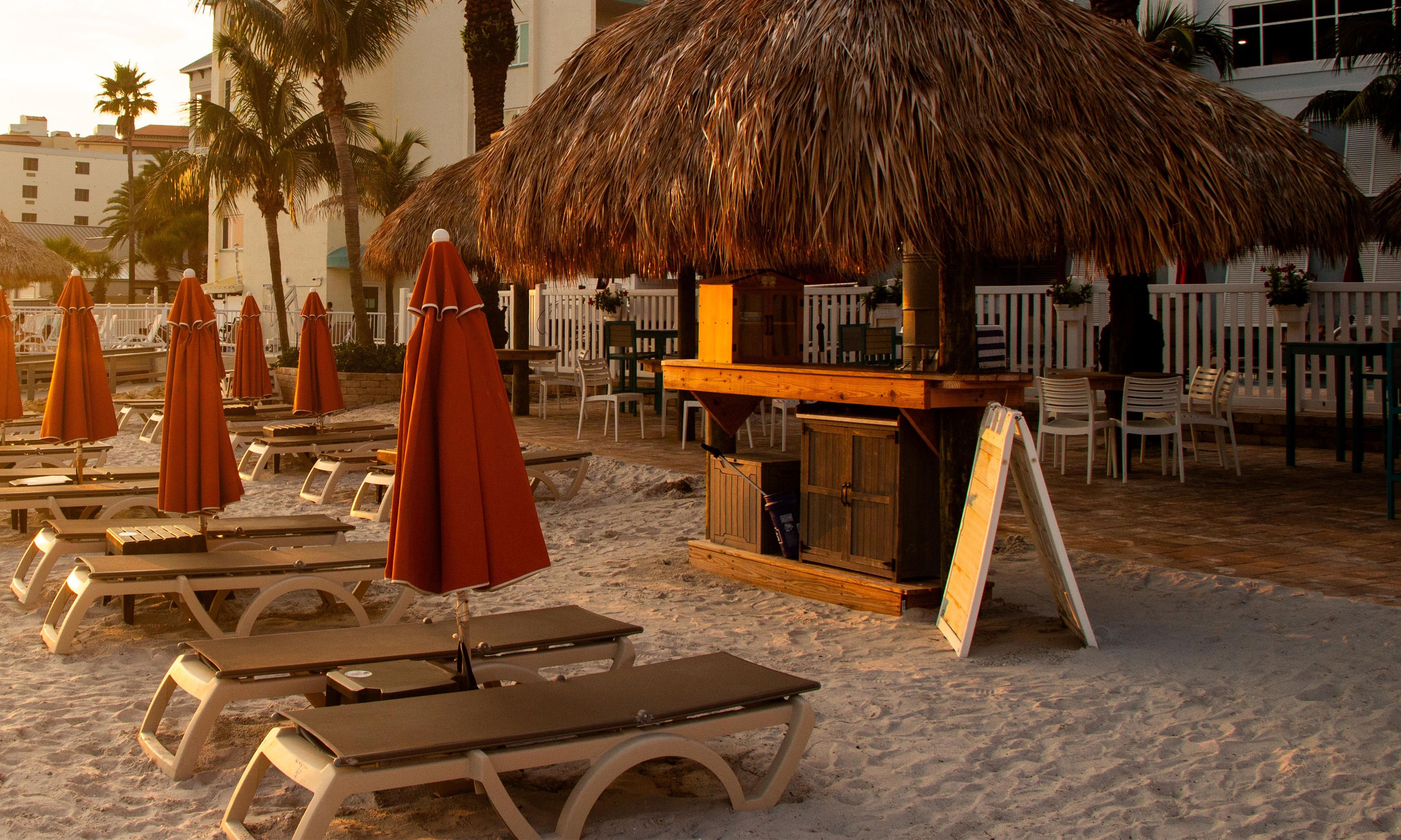 Ein Strandbereich mit Liegestühlen, Sonnenschirmen und einer strohgedeckten Struktur, wahrscheinlich eine Bar oder ein Imbissbereich.