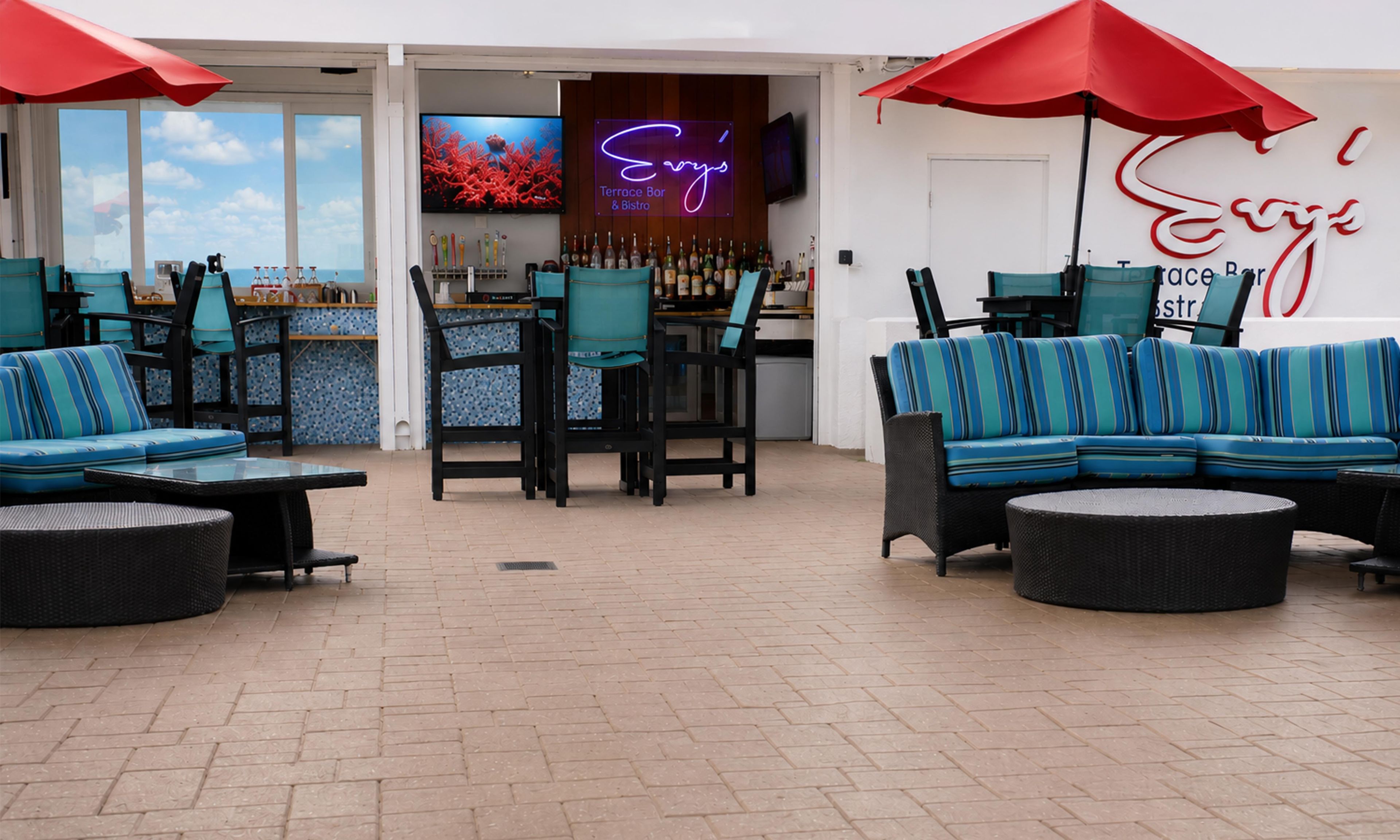 An outdoor seating area with blue-striped sofas and chairs, red umbrellas, and a bar with a neon sign.