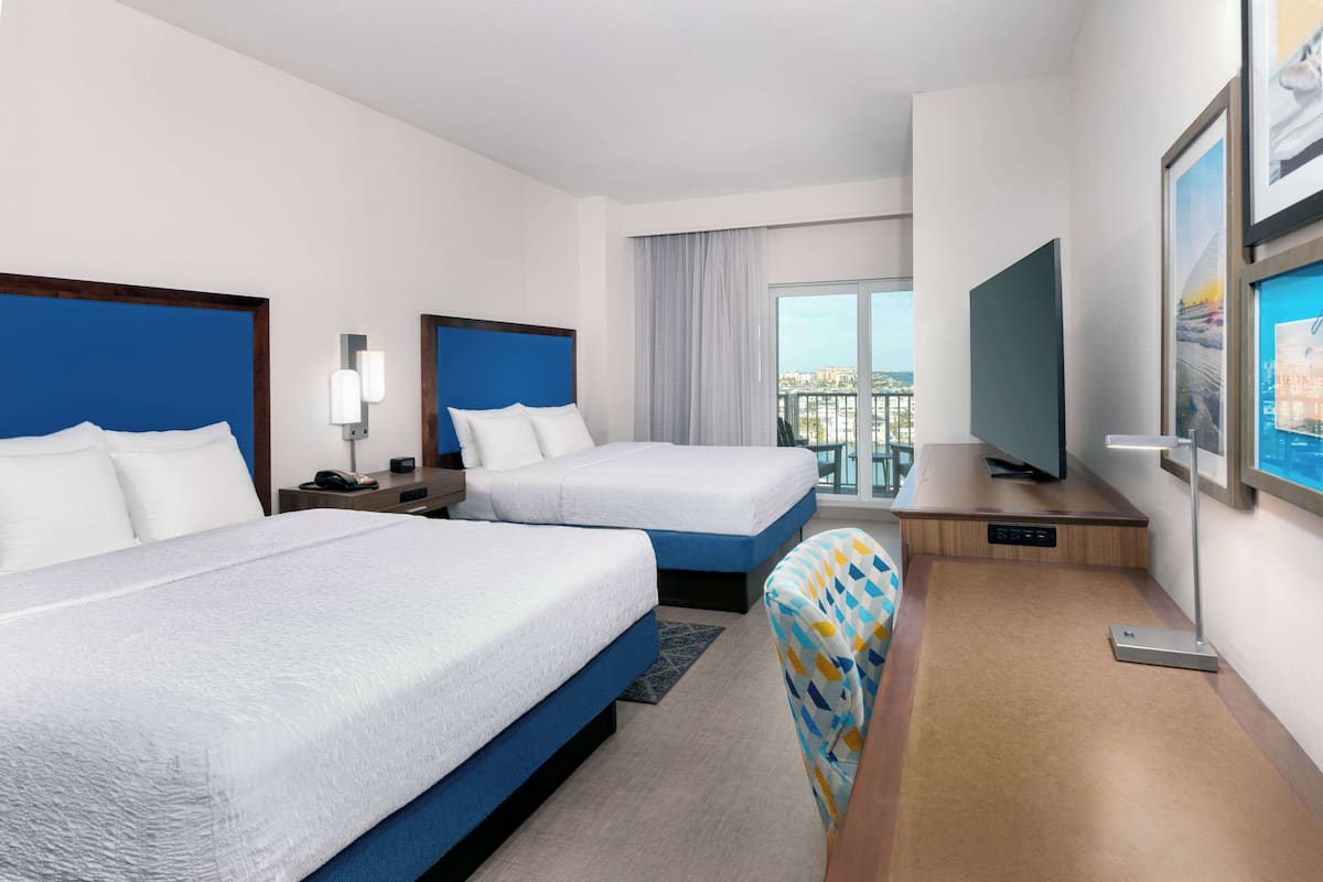 A hotel room with two beds, a desk with a television, and a large window offering a city view.
