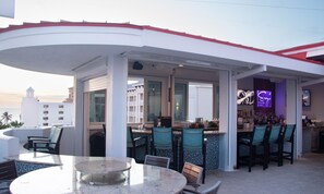 An outdoor bar area with a round table and chairs, a bar counter with stools, and a view of a building with balconies. - Hampton Inn & Suites Clearwater Beach (Clearwater Beach)