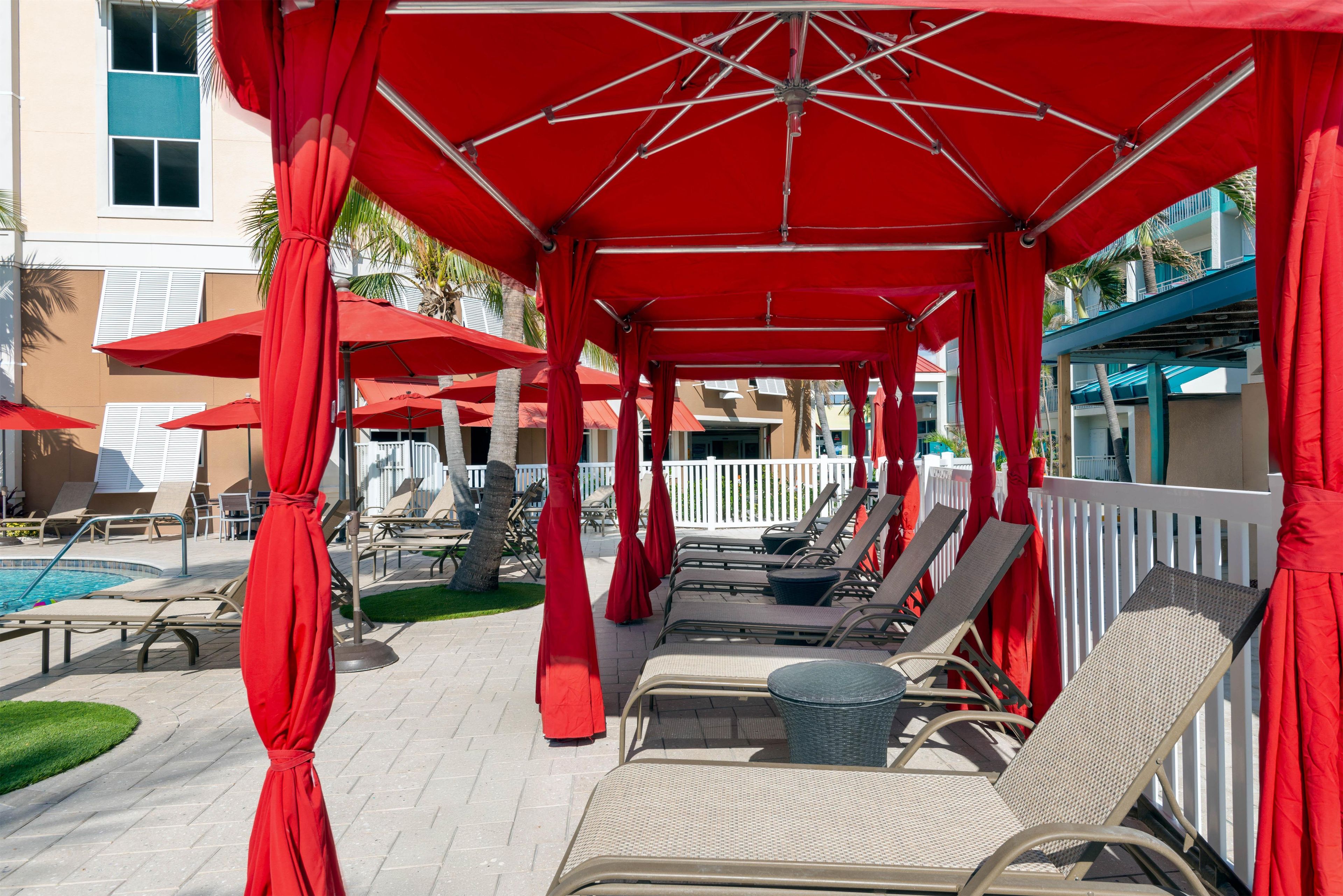 Un área de estar al aire libre con techo, sombrillas rojas, sillones reclinables y una piscina.