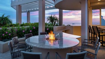 An outdoor patio with a round table and chairs, a fire feature, and a view of the ocean.