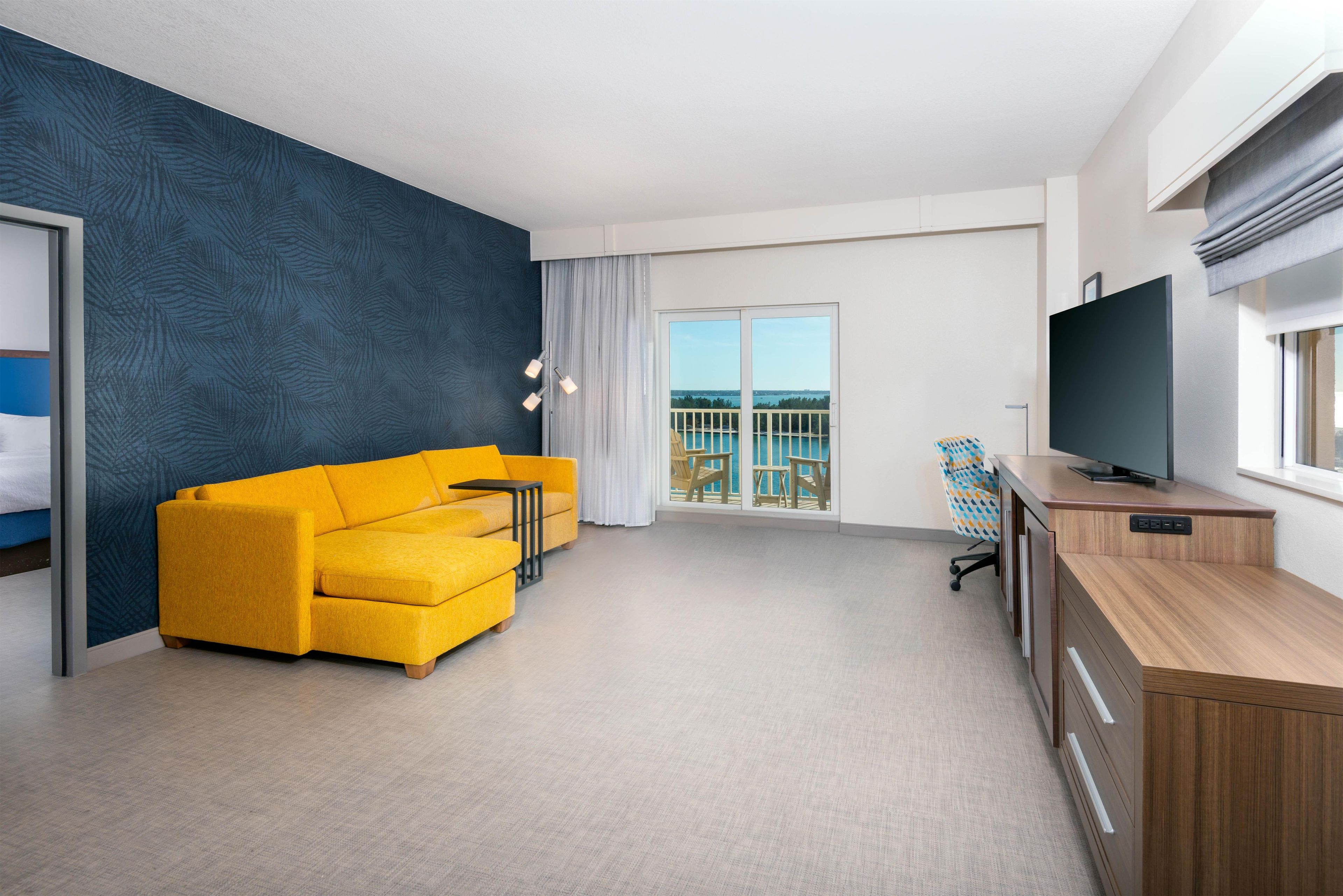 A modern hotel room with a yellow sofa, a TV, a balcony with a view of the sea, and a large window.