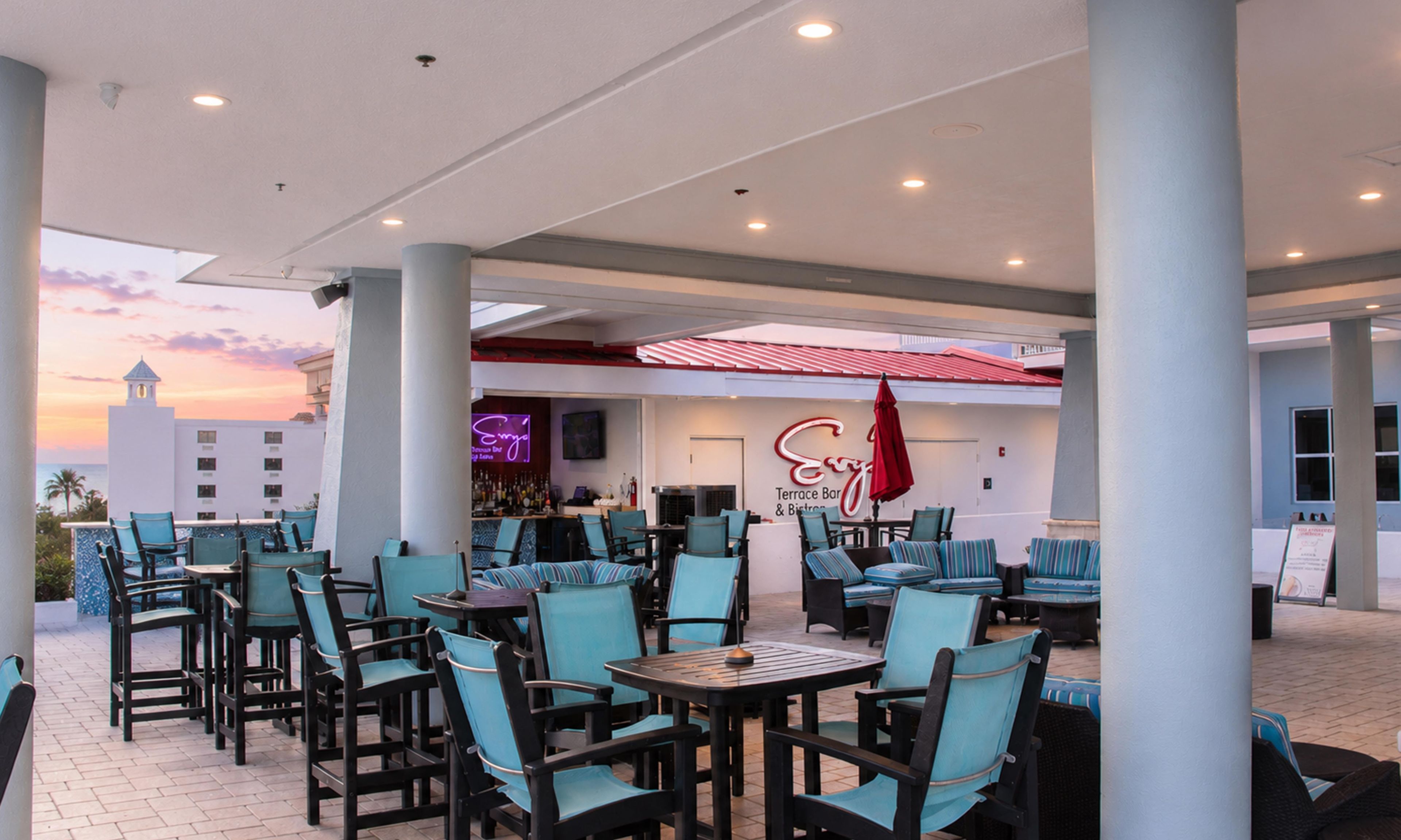 An outdoor seating area with tables and chairs, a bar, and a neon sign.