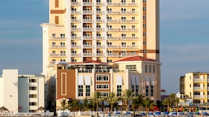 A multi-story hotel with balconies, located near a beach with palm trees and umbrellas.