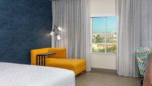 A hotel room with a bed, a yellow armchair, a small table, a view of the city, and a patterned wall. - Hampton Inn & Suites Clearwater Beach (Clearwater Beach)