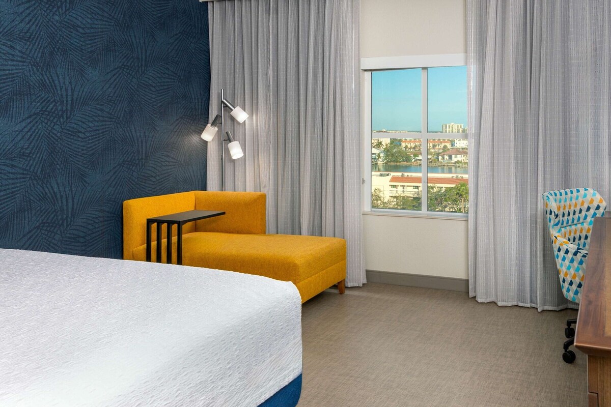 Ein Hotelzimmer mit Bett, gelbem Sessel, kleinem Tisch, Blick auf die Stadt und einer Wand mit Muster.