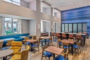 A hotel dining area with multiple tables and chairs, a blue cushioned seating area, and large windows offering a view. - Hampton Inn & Suites Clearwater Beach (Clearwater Beach)