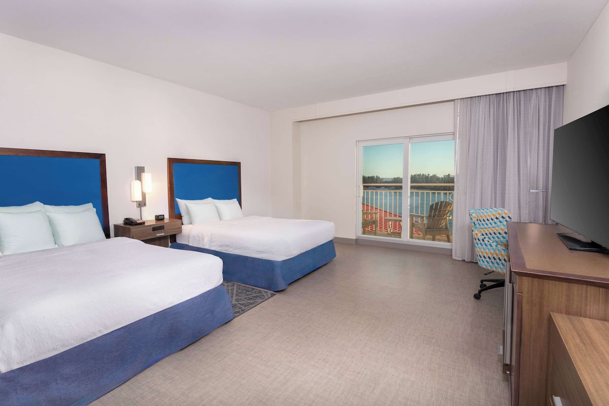 A hotel room with two beds, a desk, a TV, and a balcony with a view of the city.