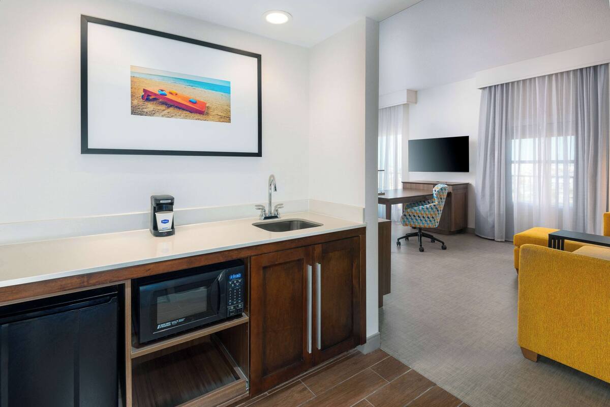 A modern hotel room with a kitchenette, a flat-screen TV, a comfortable yellow sofa, and a large window with sheer curtains.