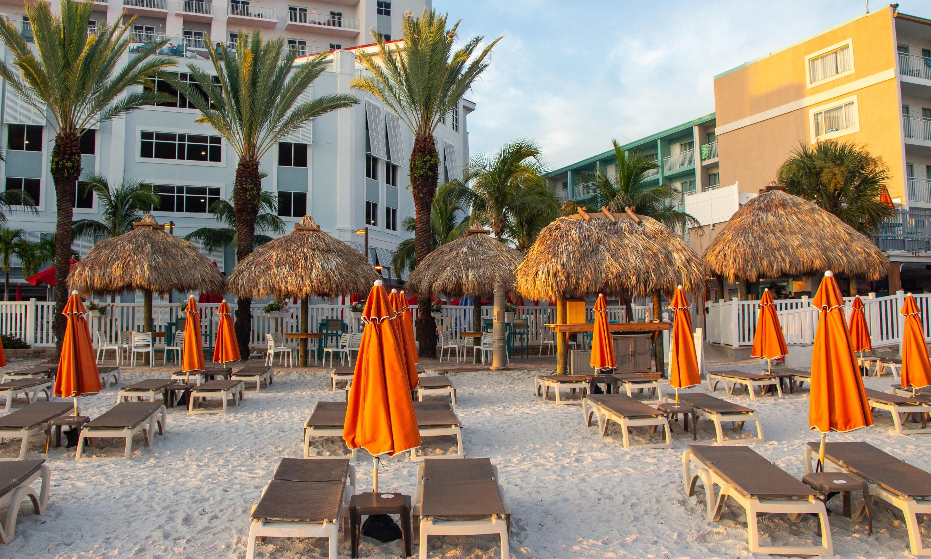 A beachside area with sun loungers and thatched umbrellas, palm trees, and a white fence.
