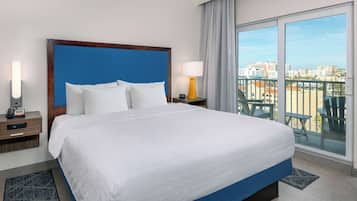 Ein Hotelzimmer mit einem großen Bett, einem Nachttisch mit Lampe, Blick auf die Stadt durch große Fenster und einem Balkon mit Sitzgelegenheiten.