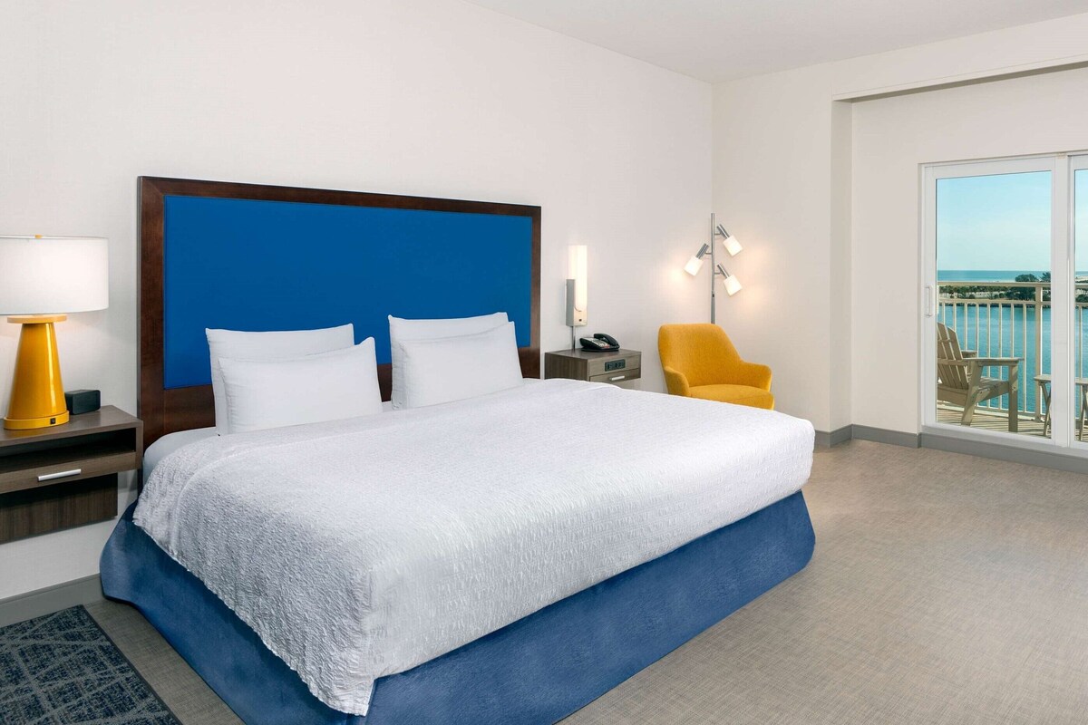 Ein Hotelzimmer mit einem großen Bett, einem Nachttisch mit Lampe, einem Sessel, einem Beistelltisch mit Telefon und einem Balkon mit Blick.
