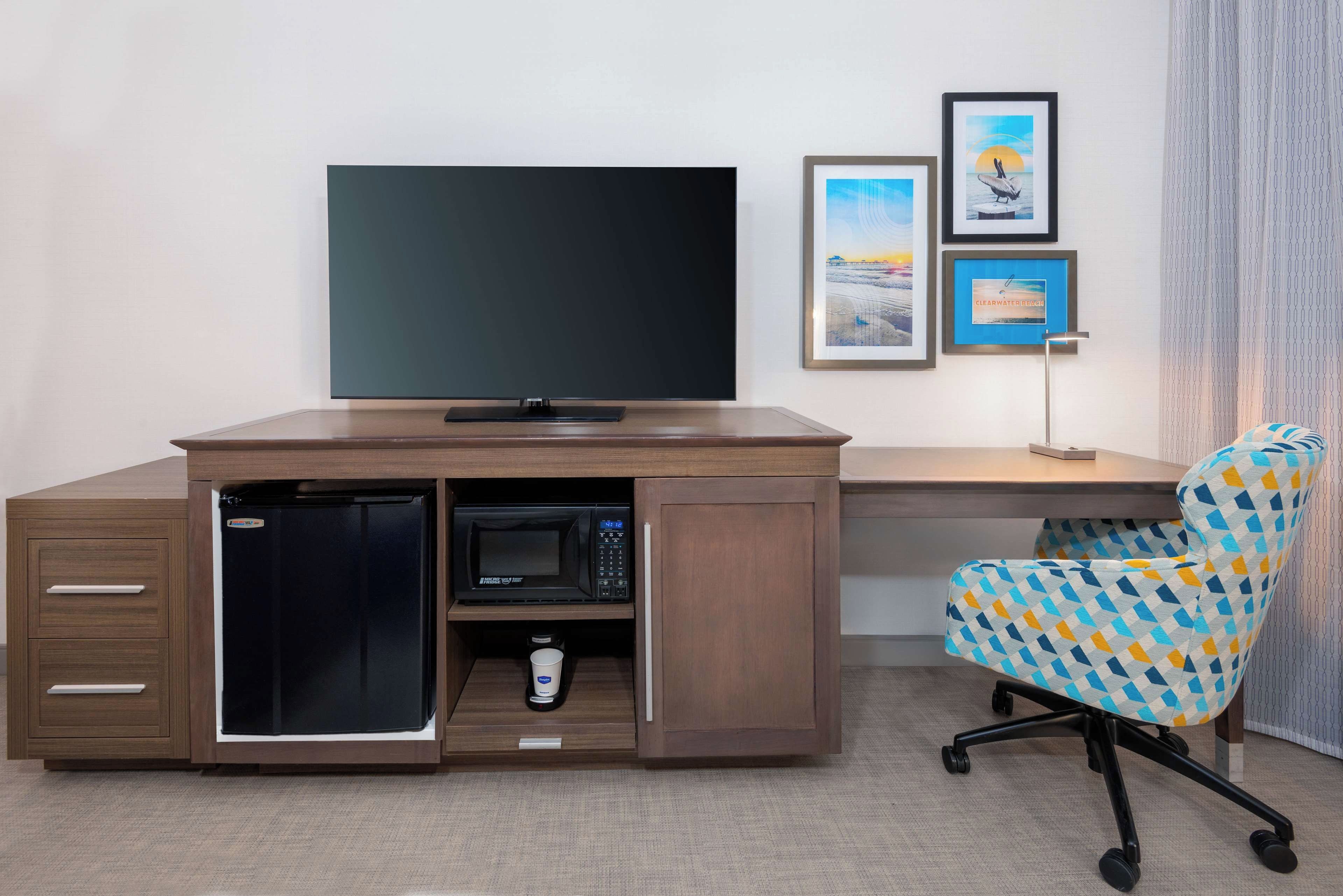 A compact hotel room with a built-in desk, a television, a microwave, and a small refrigerator. There is a chair with a geometric pattern and a framed picture on the wall.