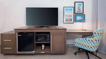 A compact hotel room with a built-in desk, a television, a microwave, and a small refrigerator. There is a chair with a geometric pattern and a framed picture on the wall.