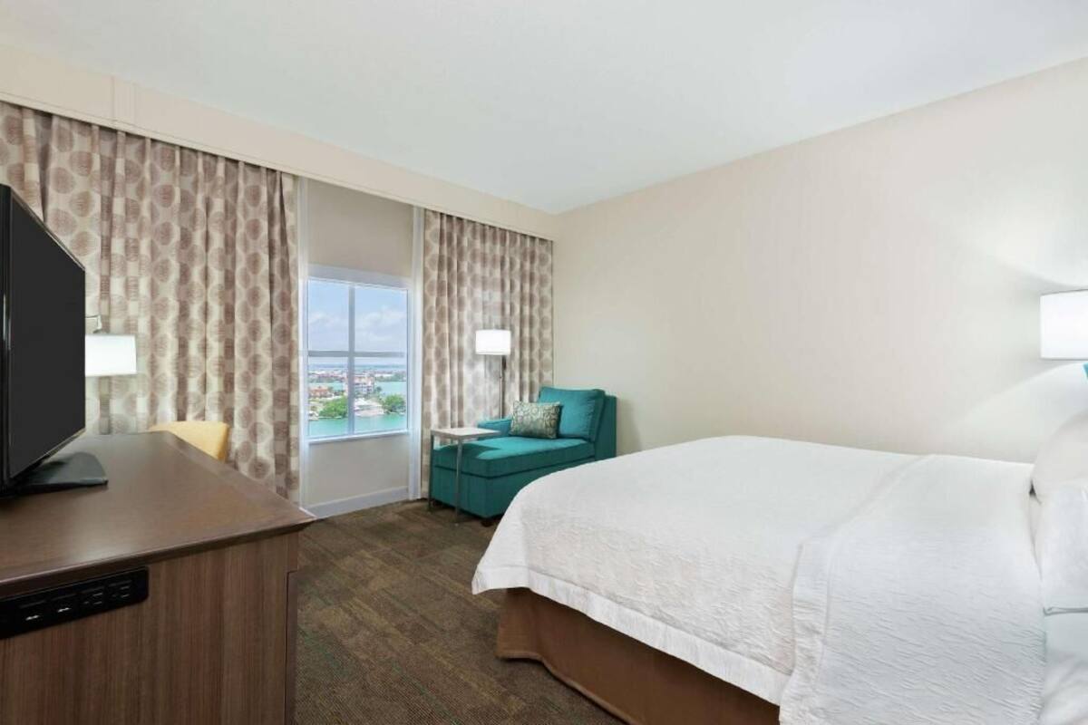 A hotel room with a bed, a desk with a television, a window with curtains, and a view of the city.