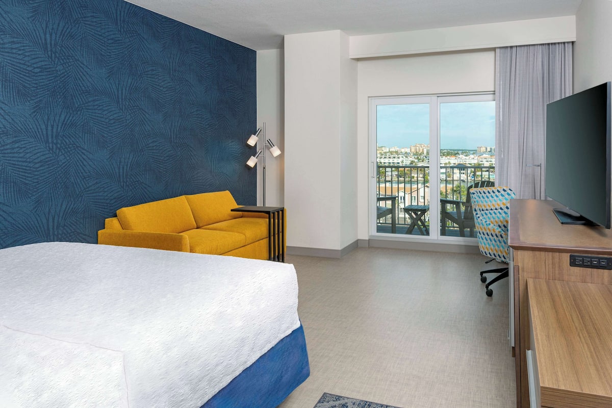 A hotel room with a large bed, a yellow sofa, a desk with a TV, and a balcony with a city view.