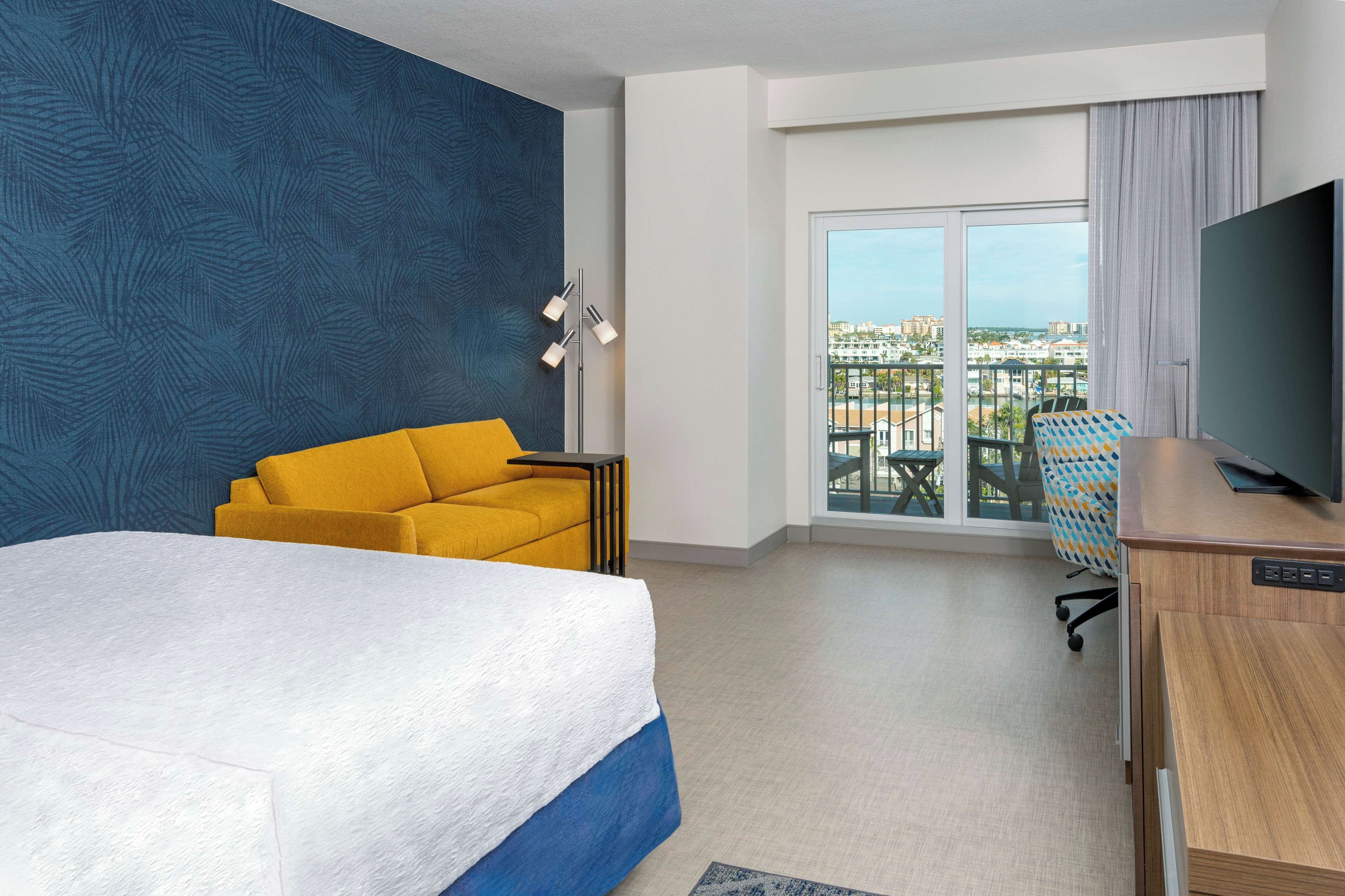 Ein Hotelzimmer mit einem großen Bett, einem gelben Sofa, einem Schreibtisch mit Fernseher und einem Balkon mit Blick auf die Stadt.