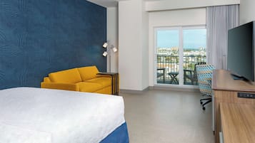Ein Hotelzimmer mit einem großen Bett, einem gelben Sofa, einem Schreibtisch mit Fernseher und einem Balkon mit Blick auf die Stadt.