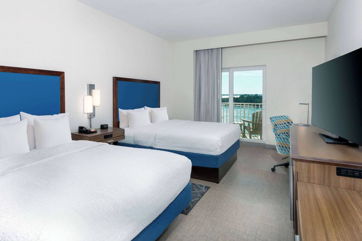 A hotel room with two beds, a desk, a television, and a balcony with a view of the water.