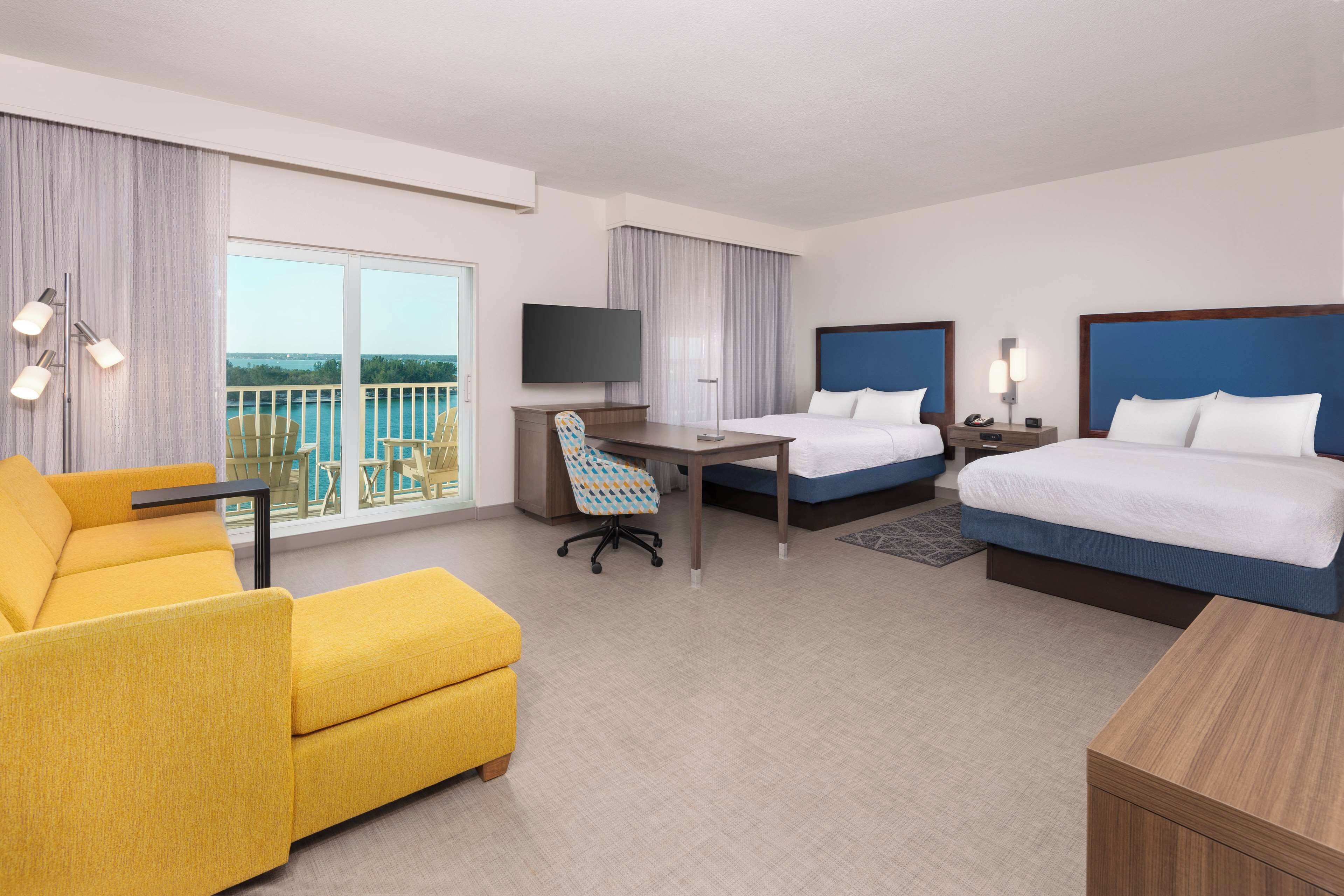 A hotel room with a balcony, a desk, two beds, a sofa, and a TV.